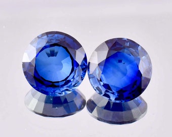 May include: Two round, faceted, blue sapphire gemstones. The sapphires are set against a white background.