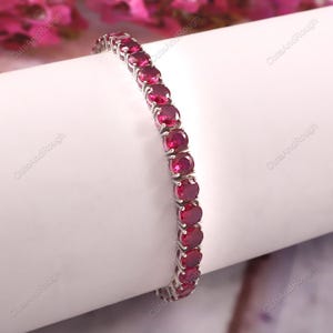 Puede incluir: Una pulsera de plata con una serie de piedras preciosas rojas oscuras de corte ovalado. Las piedras están engastadas juntas, creando una línea continua de color. La pulsera se muestra sobre una superficie blanca, con un fondo floral borroso.