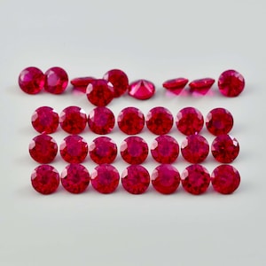May include: A collection of 25 round, red ruby gemstones. The gemstones are arranged in rows on a white surface.