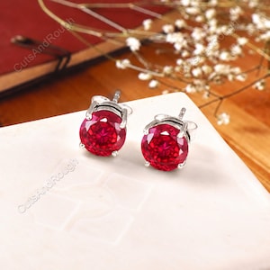 May include: A pair of round-cut red gemstone stud earrings set in silver metal prongs. Each earring features a faceted, vibrant red stone secured by a four-prong setting, showcasing a brilliant sparkle and a classic, circular design profile.