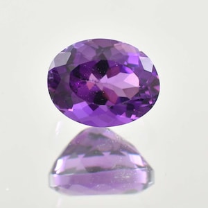 May include: An oval-cut, faceted amethyst gemstone. The gem is a deep purple color with a reflective surface, and is displayed against a white background. The facets create a play of light and shadow, enhancing its brilliance.