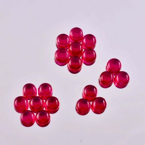 Może przedstawiać: Układ kilku małych, okrągłych kaboszonów w kolorze rubinowej czerwieni. Kaboszony ułożone są w grupy, z których niektóre tworzą wzory przypominające kwiaty. Błyszcząca powierzchnia kaboszonów odbija światło, tworząc żywą ekspozycję. Mogą być używane do tworzenia biżuterii lub innych projektów rzemieślniczych.