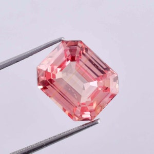 May include: A pink gemstone with a rectangular emerald cut. The gemstone is held in tweezers against a white background.