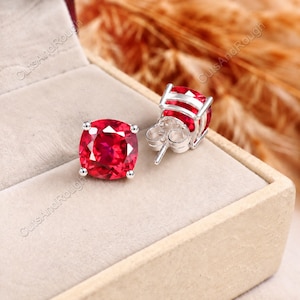 May include: A pair of cushion-cut red gemstone stud earrings set in silver-toned metal. One earring is displayed face-up showing the faceted surface, while the other is positioned on its side, revealing the silver post and butterfly clutch backing.