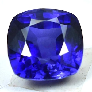 May include: A deep blue sapphire gemstone with a cushion cut. The gemstone is faceted and has a high polish.