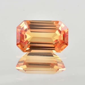 May include: An orange gemstone with an emerald cut. The gem has a rectangular shape with beveled corners and a reflective surface. The color is a warm, light orange with a hint of yellow.