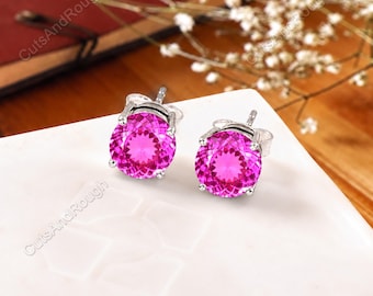 May include: A pair of stud earrings featuring round, faceted, vibrant pink gemstones set in silver-toned metal prongs. The earrings are displayed on a white surface with the text CutsAndRough visible in the corner of the image frame.