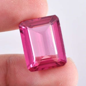 May include: A rectangular, faceted gemstone in a vibrant pink hue. The gem is held between fingers, showcasing its clarity and polished surfaces. The cut of the stone is an emerald cut, with a rectangular shape and step-cut facets.