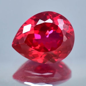 May include: A pear-shaped, deep red gemstone with a faceted cut. The gemstone is reflecting light, showing its brilliance.