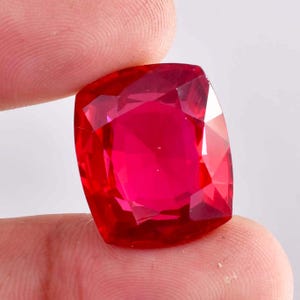 May include: A faceted, cushion-cut ruby gemstone. The gem is a vibrant red color, with multiple facets reflecting light. The gemstone is held between two fingers, showcasing its size and brilliance. The overall shape is square with rounded corners.