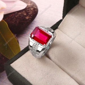 May include: A silver-toned ring featuring a large, rectangular, vibrant red gemstone set in the centre. The band is accented with two rows of small, clear, sparkling stones along the sides, creating a polished and reflective metallic finish.