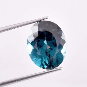 May include: An oval-cut, teal-colored gemstone held by a pair of silver tweezers. The gem has a faceted surface that reflects light, creating a sparkling effect. The stone's color transitions from a deep teal to a lighter, brighter hue.