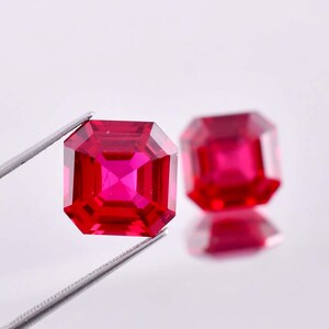 May include: Two red square-shaped gemstones with a faceted cut. The gemstones are held by tweezers against a white background.