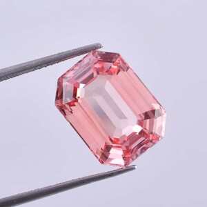 Puede incluir: Una piedra preciosa rosa de talla esmeralda sostenida por unas pinzas. La piedra preciosa es transparente y tiene un ligero tono rosado.