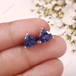 Puede incluir: Un par de pendientes de botón con piedras preciosas triangulares de color azul oscuro. Las gemas están engastadas en metal plateado. Los pendientes se muestran sobre un fondo suave y borroso, resaltando el color y la forma de las piedras.