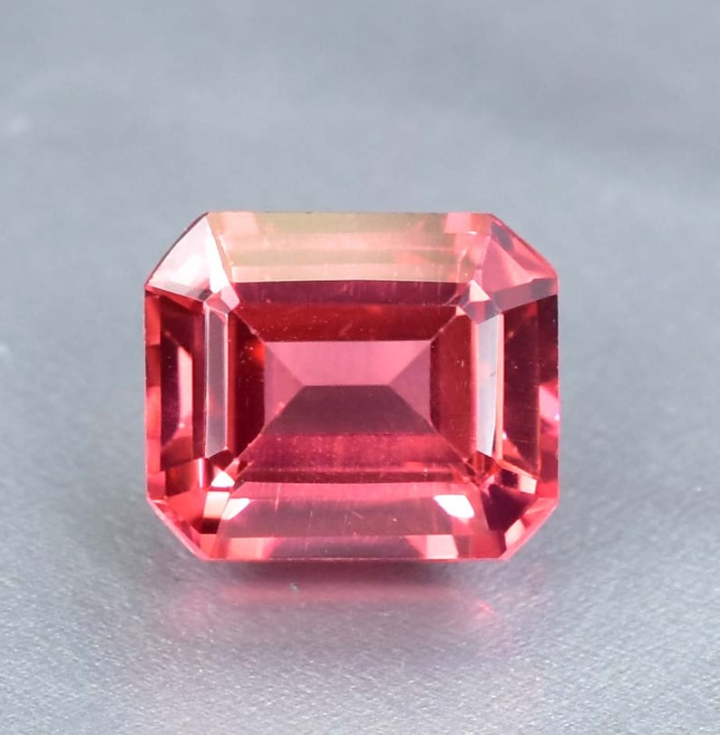 May include: A vibrant pink gemstone with a square emerald cut. The gemstone is faceted and has a high polish.
