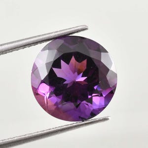 May include: A faceted, round amethyst gemstone held by tweezers. The gem displays a range of purple hues, from deep violet to lighter shades, with a star-like pattern at its center. The gem is held against a white background.