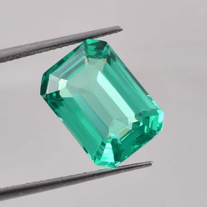 Op de afbeelding: Een levendige groene smaragd edelsteen met een rechthoekige smaragd-slijp. De edelsteen wordt vastgehouden in een pincet.