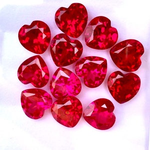 May include: A collection of twelve heart-shaped ruby gemstones, each with a deep red hue and a faceted cut. The gemstones are arranged in a circular pattern on a white surface.