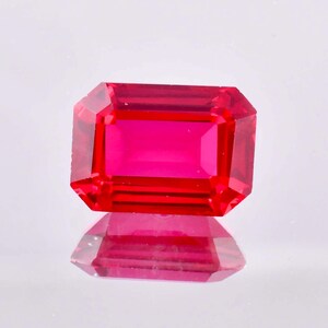 May include: A vibrant red gemstone with a rectangular emerald cut. The gemstone is faceted and reflects light.