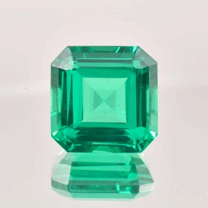 May include: A vibrant green emerald gemstone with a square cut and a polished finish. The emerald is reflecting light, showcasing its brilliance.