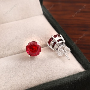 May include: A pair of ruby stud earrings. Each earring has a round, deep red gemstone set in silver-coloured metal. The earrings are displayed in a velvet-lined jewellery box.