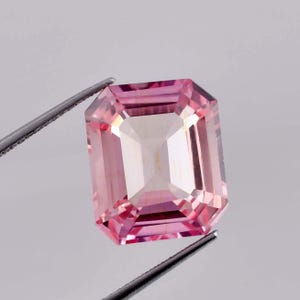 May include: A pink gemstone with a square emerald cut. The gemstone is held by tweezers against a white background.