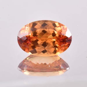 May include: An oval-cut gemstone with a warm, orange-brown hue. The gem's facets reflect light, creating a sparkling effect. The stone is set against a reflective surface, mirroring its shape and color.
