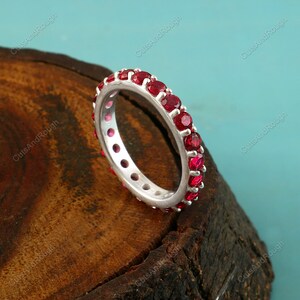 May include: A silver ring with a band of vibrant red gemstones. The ring is set against a dark wood surface with a turquoise background, highlighting the ring's color and design. A piece of jewelry.