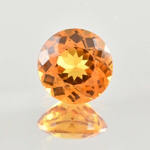 May include: A round, faceted gemstone with a brilliant orange hue. The gem's facets reflect light, creating a starburst pattern in the center. The gem is displayed on a reflective surface, showing its mirrored image.