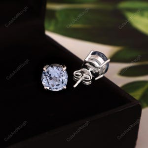 May include: A pair of light blue gemstone stud earrings. The round-cut gemstones are set in a silver metal. The earrings are displayed in a black jewelry box against a blurred green background.