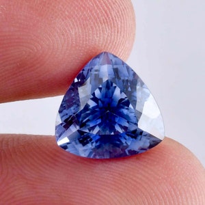 May include: A triangular, faceted gemstone with a deep blue hue. The gem is held between two fingers, showcasing its polished surfaces and intricate internal patterns. The gem's shape is a perfect equilateral triangle.