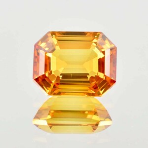 Puede incluir: Una radiante gema de talla esmeralda con un vibrante tono amarillo-naranja. Las facetas de la gema reflejan la luz, creando un brillo deslumbrante. La gema se encuentra sobre un fondo blanco, con su reflejo visible debajo.