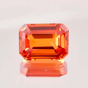 May include: A vibrant orange gemstone with a rectangular emerald cut. The gemstone is faceted and has a high polish.