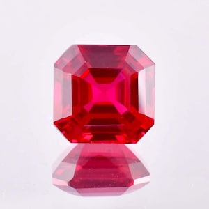 May include: A vibrant red gemstone with a square emerald cut. The gemstone is faceted and has a high polish.