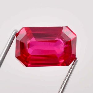 May include: A vibrant, deep red gemstone with a rectangular emerald cut. The gemstone is held in place by a pair of tweezers.
