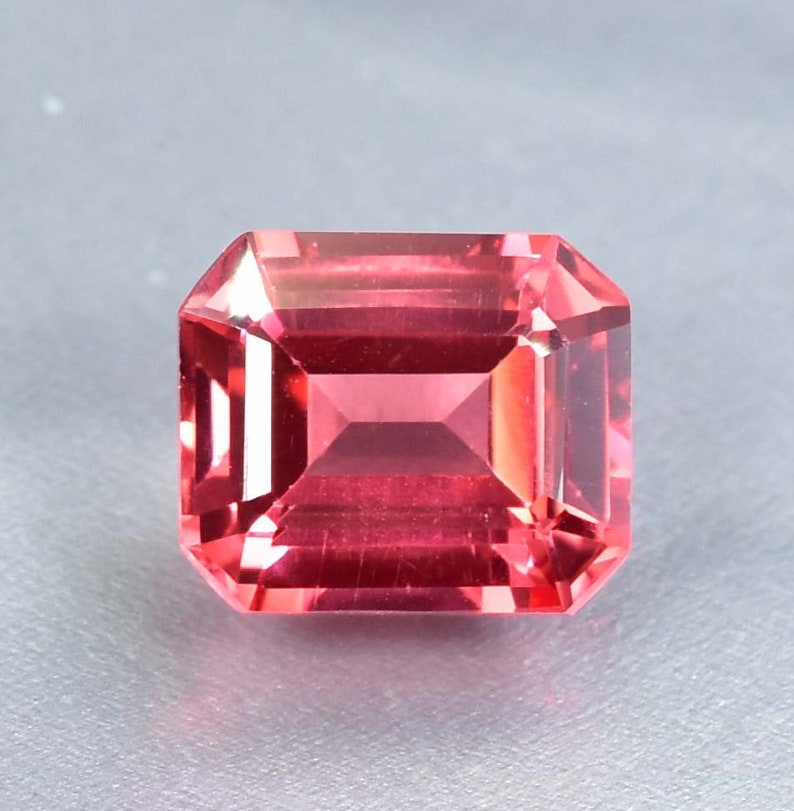 May include: A vibrant pink gemstone with a square emerald cut. The gemstone is faceted and has a high polish.