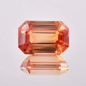 May include: An orange gemstone with an emerald cut. The gem has a rectangular shape with cut corners and facets that reflect light. The gem is sitting on a reflective surface, creating a mirror image.