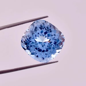 Pode incluir: Uma gema de água-marinha azul claro com um corte de almofada. A gema é segurada por pinças.