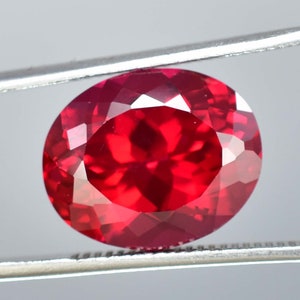 May include: A deep red oval-shaped gemstone with a faceted cut. The gemstone is held in place by a pair of tweezers.