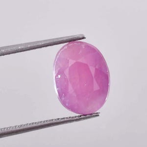 Könnte beinhalten: Ein oval geschliffener, rosa Edelstein, der von einer silbernen Pinzette gehalten wird. Die facettierte Oberfläche des Edelsteins reflektiert das Licht. Der Edelstein hat einen leuchtenden Rosaton und ein poliertes Aussehen. Die Pinzette hält den Stein vor weißem Hintergrund.