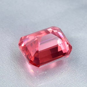 May include: A vibrant pink gemstone with a rectangular emerald cut. The gemstone is faceted and has a high polish.