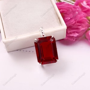 May include: A rectangular, deep red gemstone pendant set in a silver-toned metal prong setting. The pendant features a faceted cut that reflects light, with a small silver-toned bail at the top for attachment to a chain or cord.