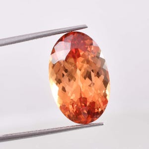 May include: An oval-cut gemstone with a vibrant blend of orange and red hues. The faceted surface reflects light, creating a sparkling effect. Held by silver tweezers, the gem is showcased against a white background.