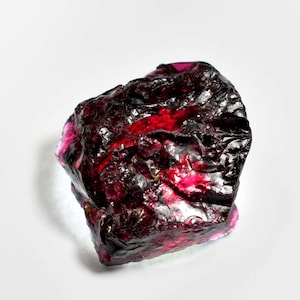 May include: A deep red, rough-cut gemstone with a dark, almost black, appearance. The gem has a natural, unpolished look.