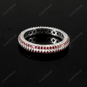 May include: A silver ring with a double row of small, round, red gemstones and a single row of clear gemstones. The ring has a detailed, openwork design on the band. The ring is set against a black background.