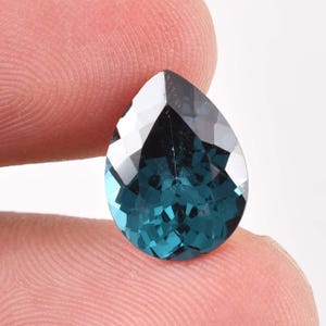 May include: A faceted, teardrop-shaped gemstone with a deep teal color. The gem has a polished surface with multiple facets that reflect light, creating a sparkling effect. The gem is held between two fingers against a white background.