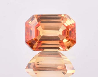 May include: A faceted, rectangular gemstone with an orange-yellow hue. The gem is cut with multiple facets, reflecting light and creating a brilliant appearance. The gemstone is displayed on a reflective surface, showing its mirrored image.