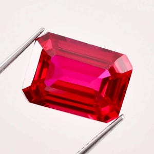 May include: A vibrant, rectangular-cut ruby gemstone, held by silver tweezers. The gem displays a deep red hue with facets that catch the light, showcasing its clarity and brilliance. The ruby is a symbol of passion and luxury.