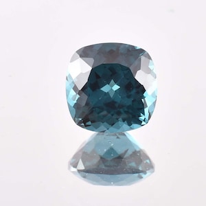 May include: A cushion-cut gemstone with a teal and blue color gradient. The gem is highly reflective, with many facets catching the light. The stone is set against a white background, with its reflection visible below.